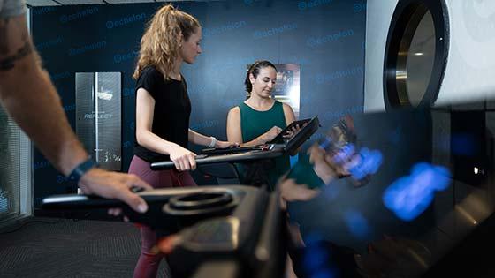 A woman with a high curly ponytail in exercise attire walking on an Echelon treadmill while another woman with dark hair and a green shirt observes and tracks progress on a smart device.
