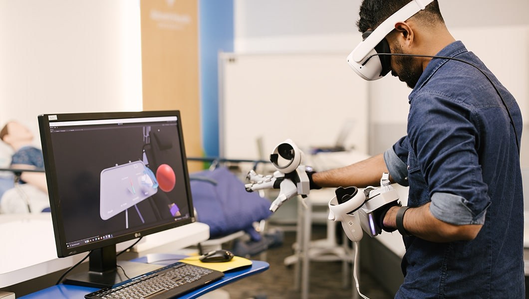 A man in a navy blue button down shirt wearing virtual reality gear on his head and heads while interacting with a screen in front of him.