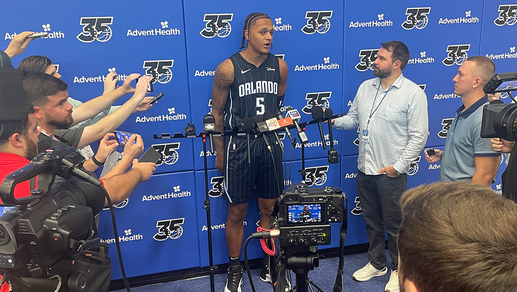 A basketball player in uniform stands against an Orlando Magic step and repeat while speaking to the press.