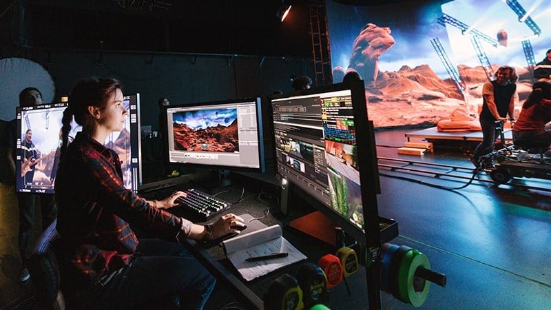 A student sits at computer monitors in a virtual production studio. They are using Unreal Engine to modify the film set.