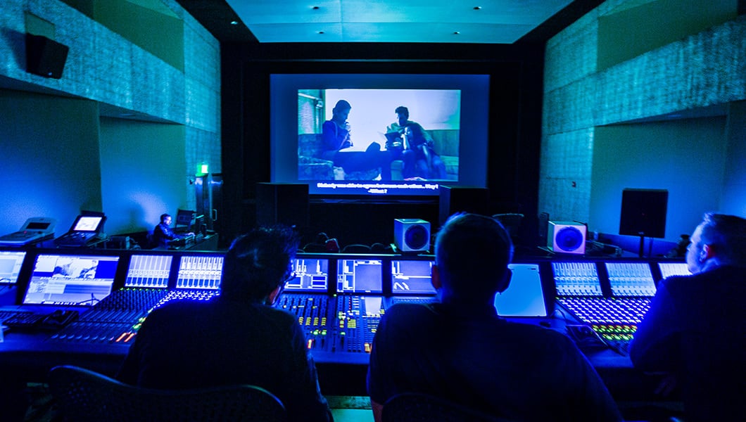 Four sound engineers sit behind an audio mixing console in the Dub Stage. The room is dark and a movie is playing on a projection screen.