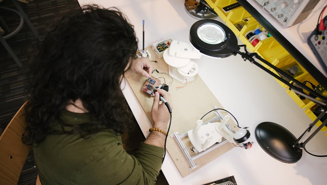 A student sits at a desk and works on a circuit board. Various circuitry parts lay around the desk as well as a lamp.