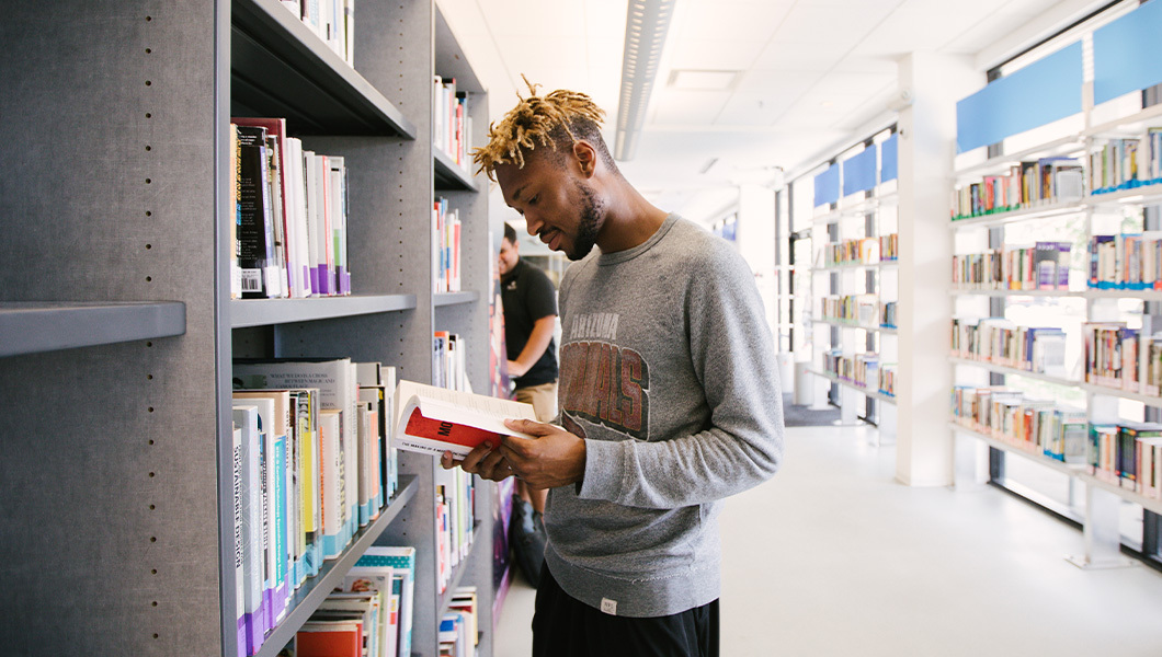 A college student reads a book in a sunny aisle lined with shelves in a quiet, modern library setting.