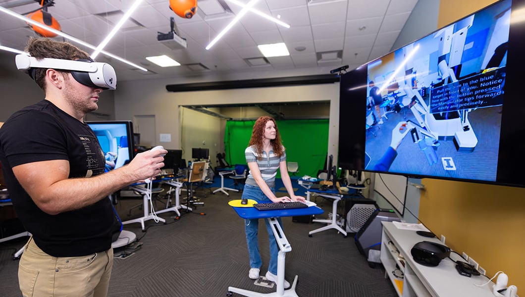 Two students stand in a classroom facing a screen. One student wears a VR headset and holds a controller while the other student watches the screen
