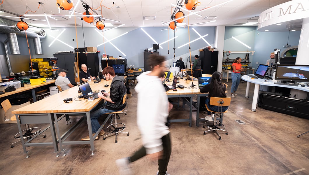 Students and instructors sit at tables, work on fabricated projects, and use a 3D printer in the brightly lit Fab Lab.