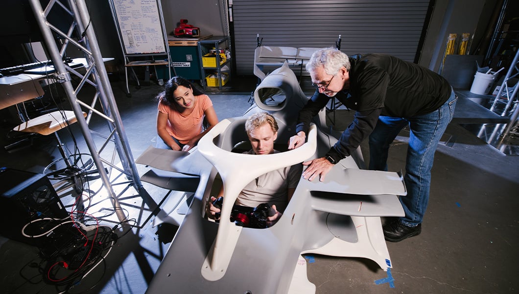 A student sits in the cockpit of a small fabricated Formula One racecar while another student and instructor look on in Full Sail’s Simulation Lab.