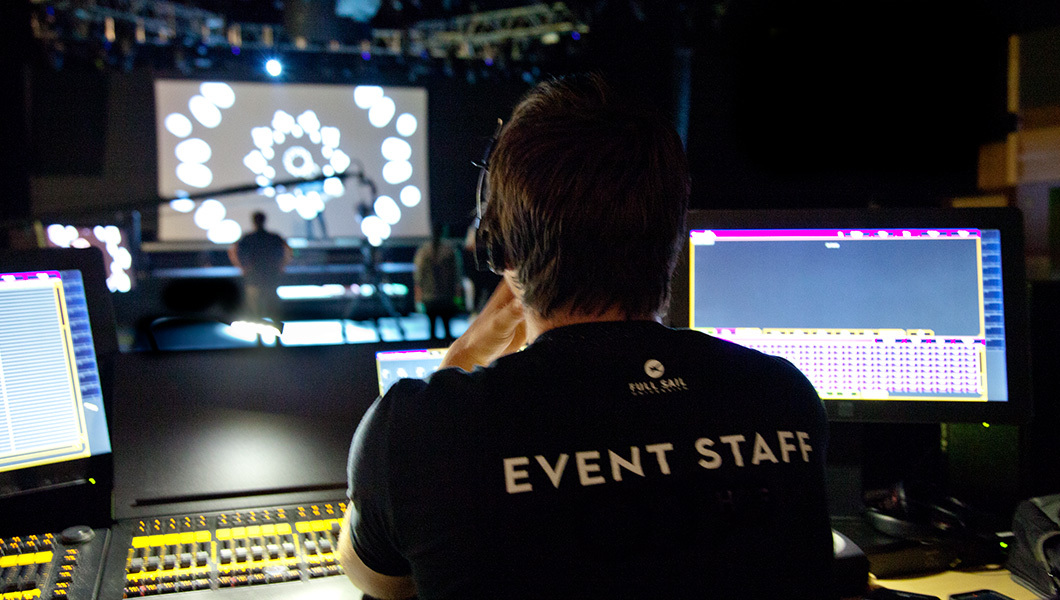 Person with 'Event Staff' on back of shirt watching monitors and stage.