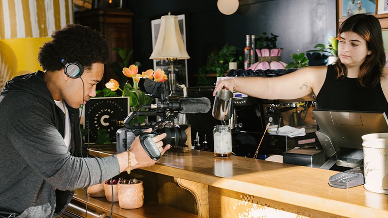 A student holds a camera and films a barista pouring a drink at a coffee shop.