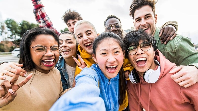A group of eight smiling young people posing for a selfie outdoors on a sunny day.