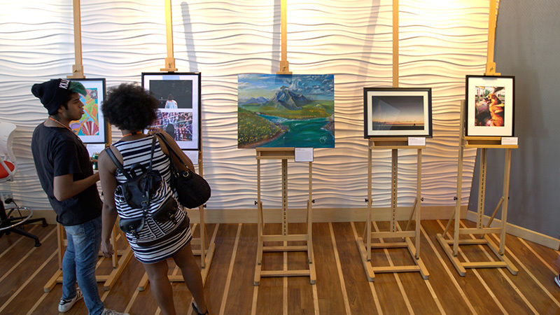 Students gather around several pieces of artwork displayed on large wooden easel stands.
