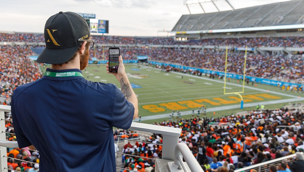 A student wearing a Full Sail baseball cap stands in the bleachers in a football stadium. They are filming the game on their cell phone.