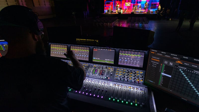 A sound engineer operates a mixing console, adjusting audio levels for a brightly lit stage performance in a dimly lit venue.