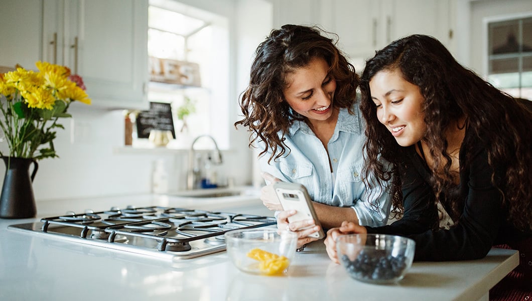 A mother and daughter standing at a kitchen island smiling together and looking at a cell phone that the mother is holding.