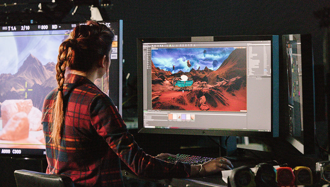 A woman sits at a desk in front of three large monitors in the Studio VI. Two of the monitors show an alien planet with red mountains and several moons.