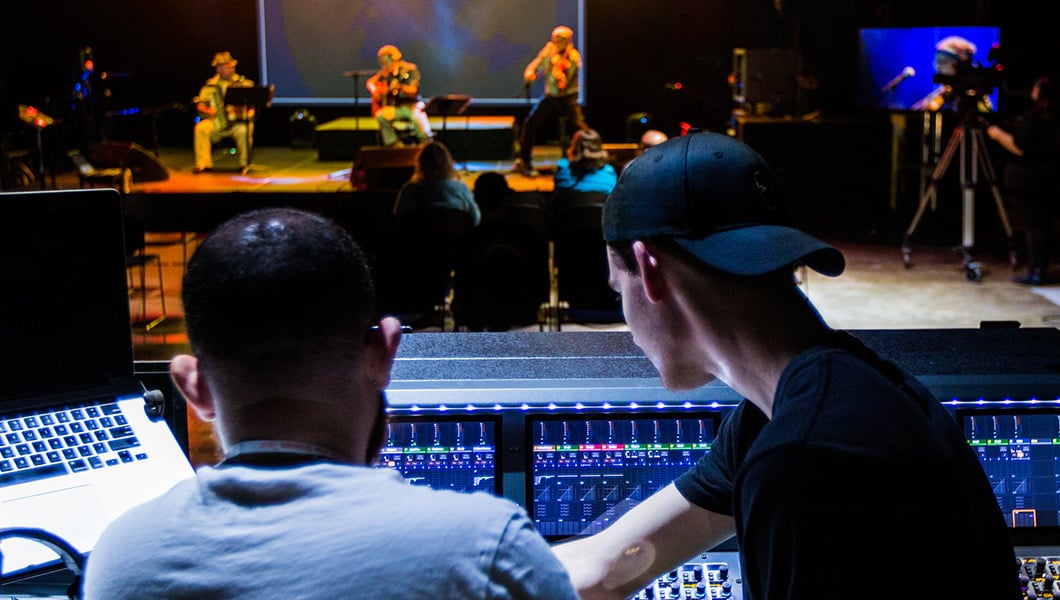 Live Event Production students work on audio, video, and lighting consoles in front of a stage. A three-person band is playing.