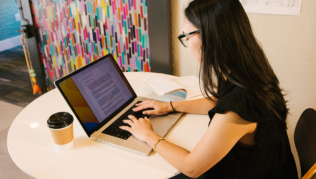 A student with long black hair and glasses sits at a table in a classroom. They are writing on a laptop and have a cup of coffee on the table.