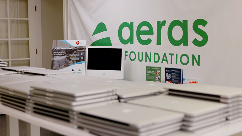 Several piles of MacBook Pro laptops sit on a table, with a backdrop featuring the Aeras Foundation logo hanging behind them.