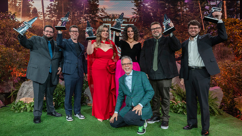 Full Sail’s 15th Hall of Fame inductees stand together in formalwear and hold their Sailees. Full Sail President Garry Jones crouches in front of them. They are on a stage designed to look like the outdoors, complete with AstroTurf and fake rocks and plants.