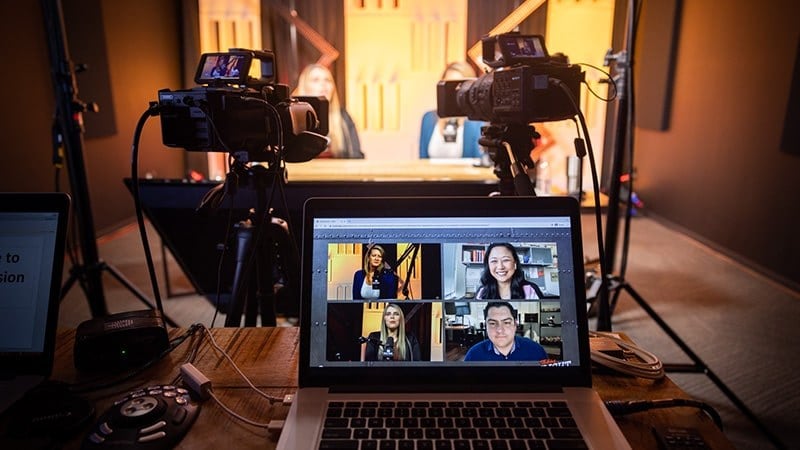 A laptop sits on a desk in a podcast studio displaying all four participants’ videos. Two cameras point at Heather Torres and Maria del Mar Herrero, who are out of focus.