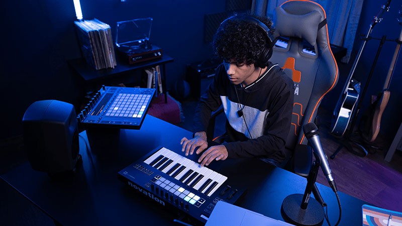 A young man with medium length curly brown hair is seated in a grey and orange Full Sail Armada gaming chair while using audio equipment at his desk.