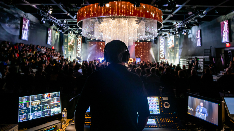 An audio professional wearing headphones sits behind an audience in Full Sail’s Fortress. They are in front of an illuminated stage.