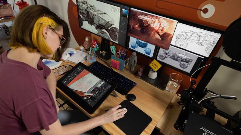 A girl with short brown hair with yellow streaks works on several drawings using an elaborate tablet and desktop computer setup.