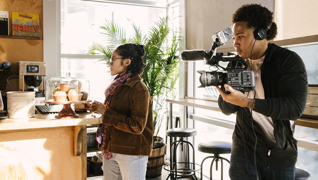 Two digital cinematography students stand in a café. One of them is holding a video camera and wearing headphones while the other looks on.