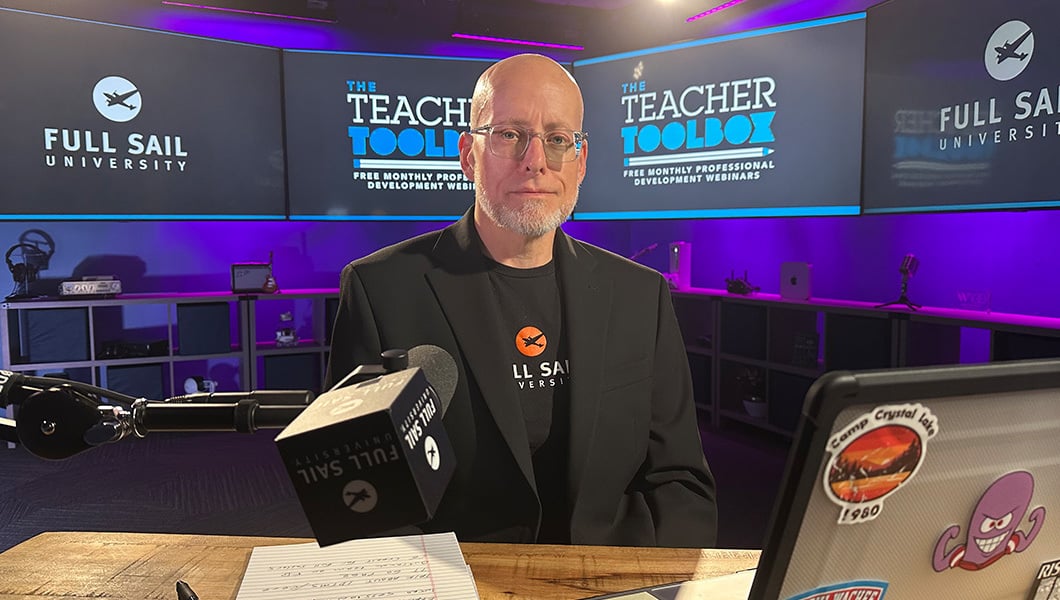 A man sits in a studio setup for Full Sail’s Teacher Toolbox webinar series, with multiple branded screens behind him.