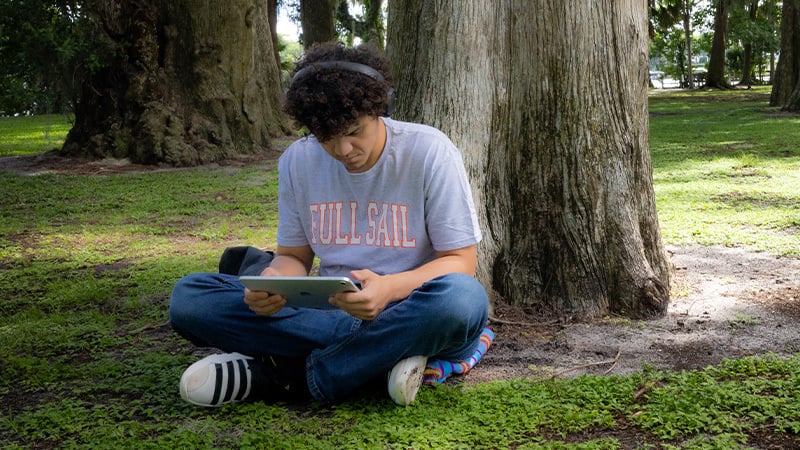 Student in a grey Full Sail shirt studies on an Apple iPad while sitting under a tree in a quiet, wooded park.