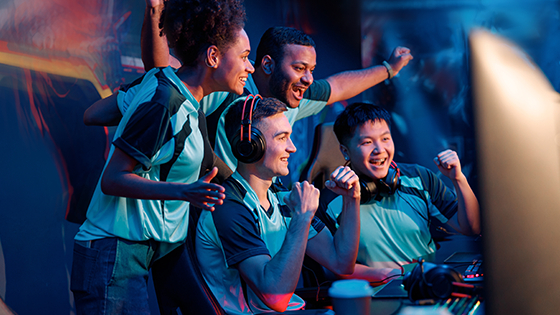 A group of three esports players cheering behind their teammate who is seated at a gaming station while wearing a headset.
