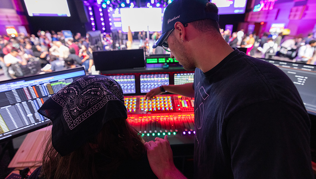 Stage production team members stand behind a desk with monitors and sound boards. One wears glasses and a baseball cap and one wears a bandana.