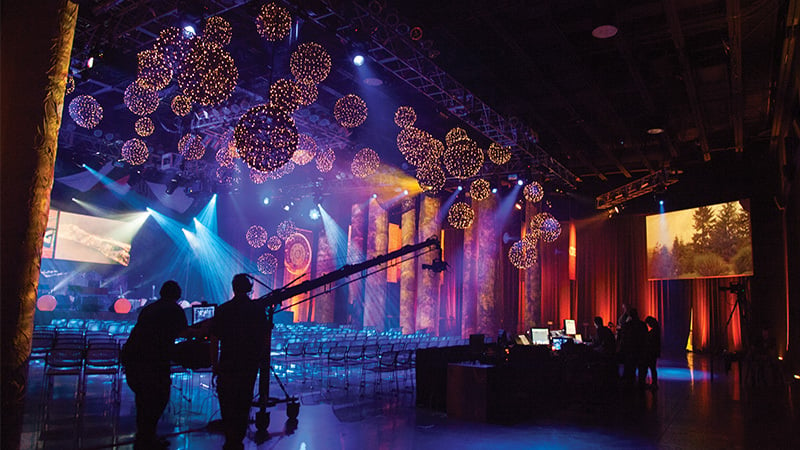 Show Production students work at computers and cameras in a large venue. Glowing orb lights hang from the ceiling and video screens hang behind the stage.