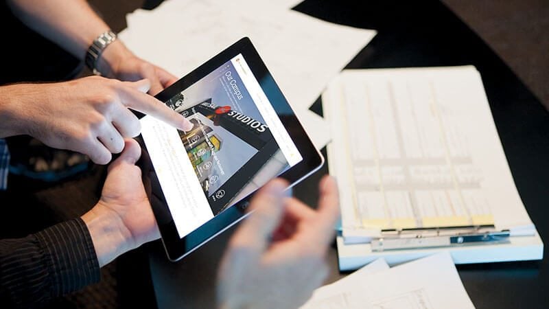 A set of hands holding a tablet while another set of hands points to the screen which displays the Full Sail University website.