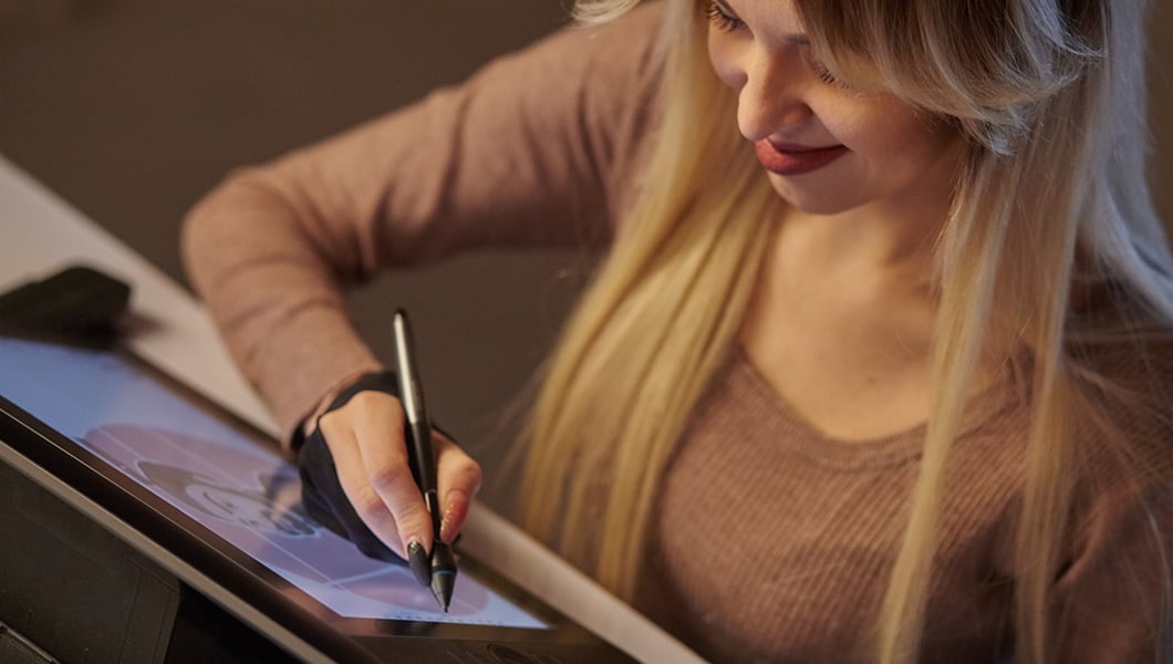 A close-up shot of a young woman with blonde hair and a brown top sketching on a tablet while using a digital pen.