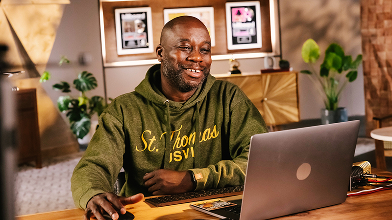 Leslie Brathwaite sits at a laptop. He is smiling and there are records and music awards in on the wall behind him.
