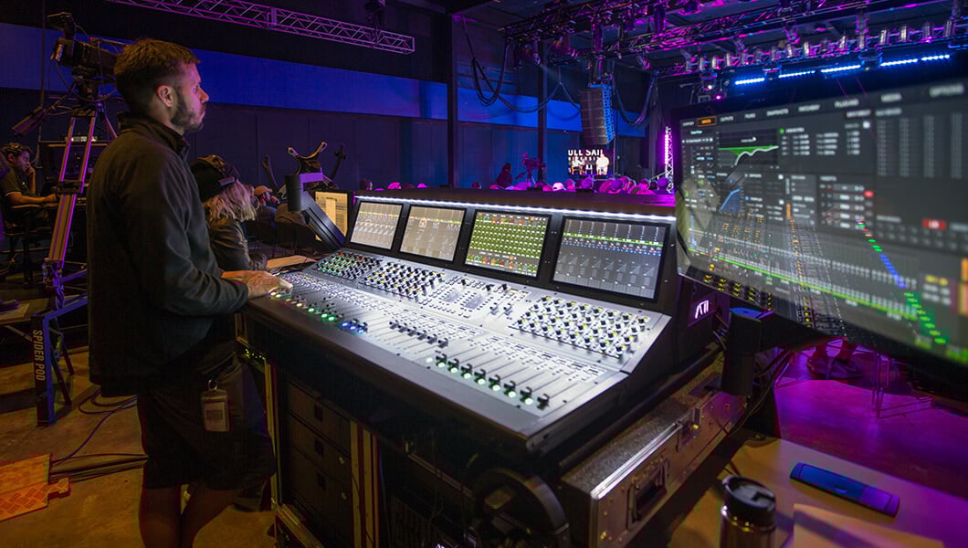 Man works behind a sound board next a large computer screen at an event.