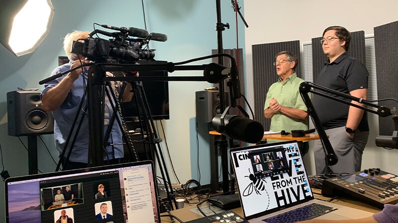 A live recording setup with two hosts on camera, surrounded by lights, microphones, and a multi-screen virtual production setup.