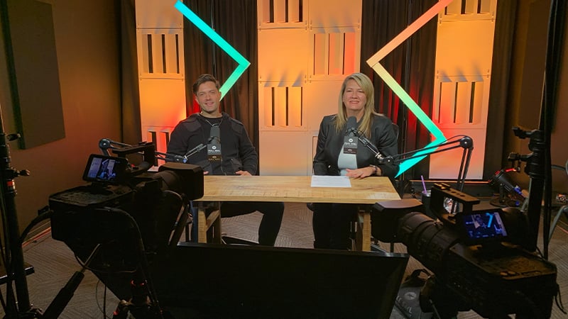 Dr. Heather Dartez and Ethan Curtis are siting in a podcast studio together, surrounding by cameras and microphones. They are both wearing black and smiling.