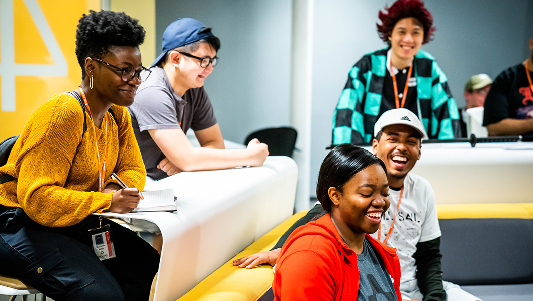 A group of Full Sail students seated around a couch in a classroom while smiling and laughing together.