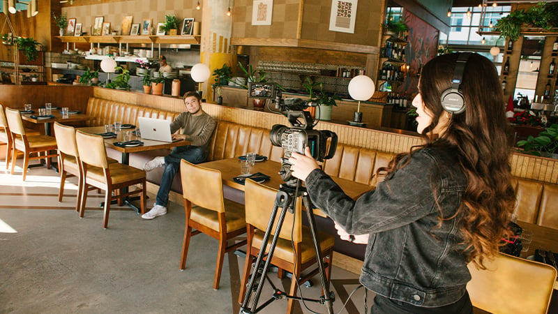 A person is operating a camera in a coffee shop while another person on a laptop is seated in view of the camera.