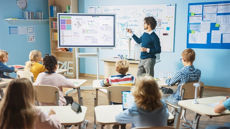 A man in the front of a light blue classroom controlling a smart board with students backs out of focus in the foreground.