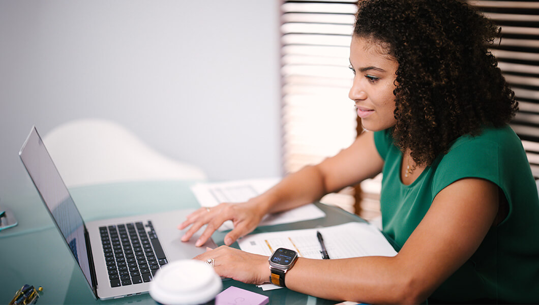 A woman in a green blouse with tight brown curly hair is seated at a desk while working on a laptop.