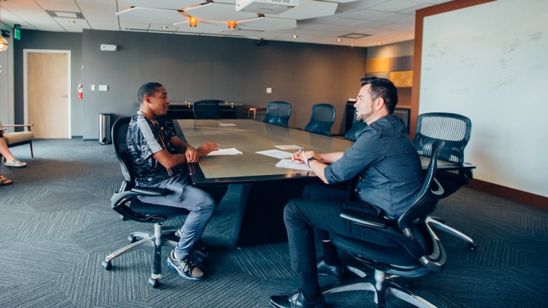 A campus mentor sits with a student and chats at a table in a Full Sail conference room. Paperwork is spread out in front of them.