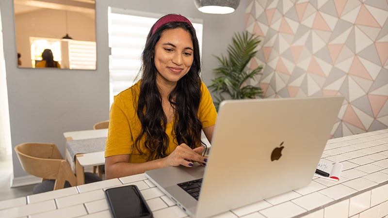 A young person with long dark hair., a yellow blouse, and a red headband is smiling and sitting at a counter while using their laptop.