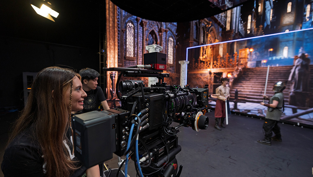 Students film a scene in front of a digital screen. Two students stand behind the camera and two students are dressed in costumes in front of the camera.