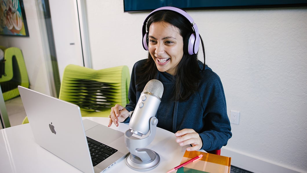 A smiling student wearing purple headphones sits at a table in a classroom. They have a laptop, a microphone, and a notebook in front of them.
