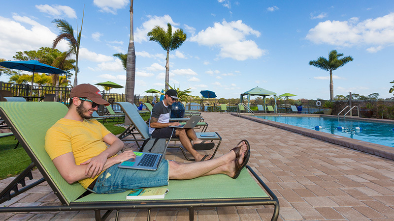 Two young men are sitting on lounge chairs while working on their laptops at a pool on a sunny day. They are both wearing sunglasses and baseball caps.