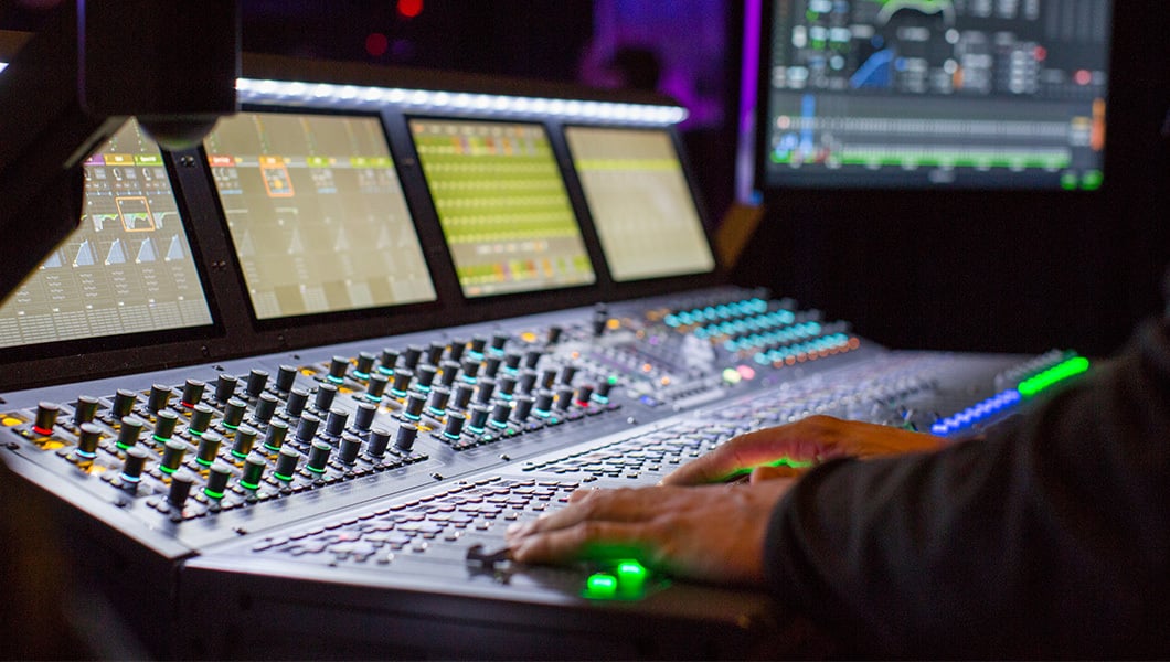 A control booth featuring several display screens and colorfully lit knobs. A pair of hands is working on the large machine.