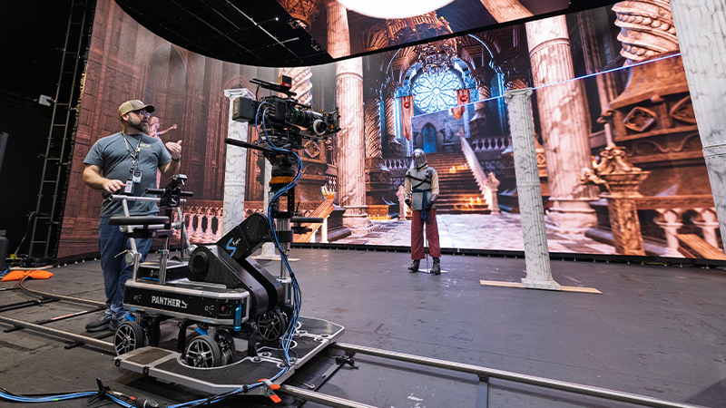 An instructor stands behind a camera dolly while an actor dressed as a knight stands in front of a virtual set featuring the interior of a castle.
