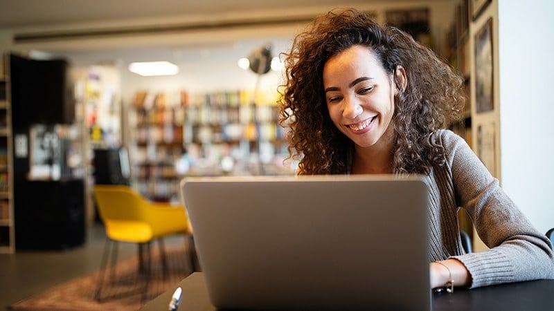 A woman with curly brown hair and a grey cardigan is seen smiling while seated in front of her laptop in a library.